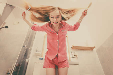 Woman wearing pajamas in bathroom having fun while brushing her long blonde hair, windblown hairdo, shot from bottom.の写真素材