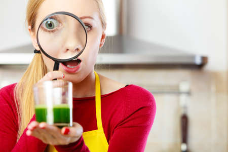 Shocked woman looking through magnifying glass at green smoothie juice made of vegetables thinking if its healthy.の写真素材