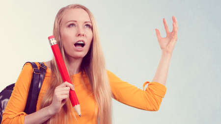 Nervous young blonde teenage girl going to school or college wearing backpack and holding big pencil feeling very stressed outの写真素材