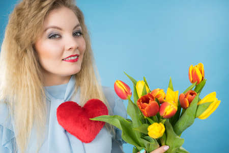 International womens or valentines day. Attractive happy woman blonde hair fashion make up holding tulips bunch and red heart sign. On blueの写真素材
