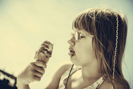 Summertime joy, summer recreation outside concept. Toddler girl in eyeglasses eating ice cream on beachの写真素材