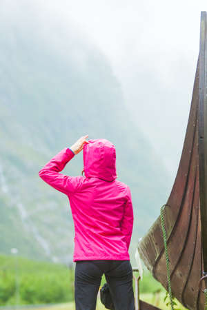 Female tourist standing near old wooden viking boat in norwegian nature, looking at misty mountains. Rainy foggy day. Tourism and traveling conceptの写真素材