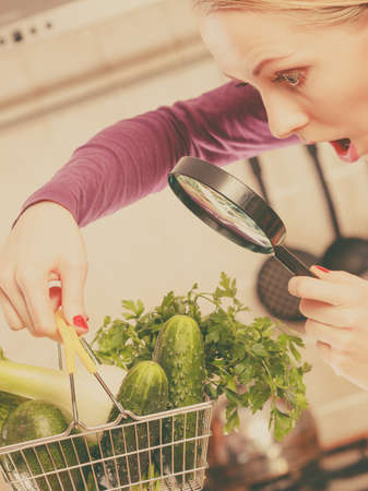 Buying healthy dieting food concept. Woman in kitchen having many green vegetables looking through magnifier at shopping basket trolley.の写真素材