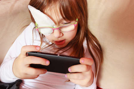 Childhood, childcare, being polite concept. Little toddler girl with bandage on eye playing games on smartphoneの写真素材