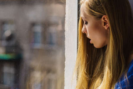 Shocked young blonde teenage woman looking through window seeing something horrible outside.の写真素材