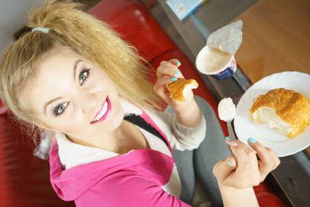 Happy cheerful young teenage woman eating croissant bun and cottage cheese yoghurt