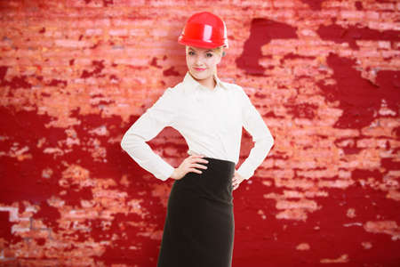 Portrait of attractive female engineer. Woman architect in red safety helmet on old brick wall...