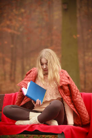 Autumn time education concept. Blonde girl with book. Attractive woman sitting on blanket in the park relaxing, foggy misty dayの写真素材