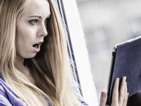 Long hair teen girl on window sill watching on tablet pc computer, reading surprising news on...