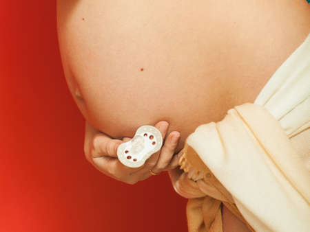 Parenthood, waiting for baby concept. Adult woman showing her big pregnant belly holding pacifier. Indoor shot on red backgroundの写真素材