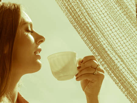 Young woman sitting on windowsill looking through window enjoying her free time, relaxing while...