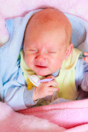 Infant care, teething pain, colic ache concept. Little newborn baby crying in bed surrounded with blankets holding teatの写真素材