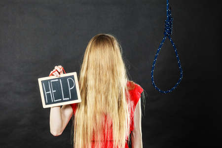 Suicidal unrecognizable skinny woman having face covered with blonde hair holding help sign on dark board next to hanging rope with knot.の写真素材