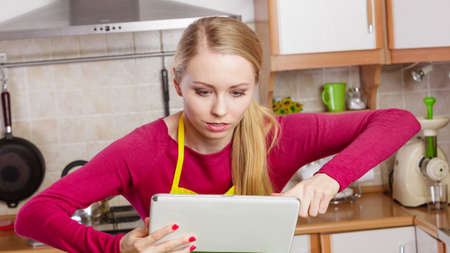 Young woman in kitchen having many green vegetables on table, holding tablet thinking about cooking something and searching for recipes in internetの写真素材