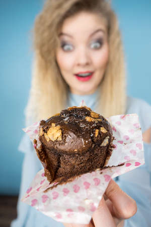 Sweet food sugar make us happy. Attractive blonde woman holds yummy chocolate cupcake in hand, open mouth, craving for cake. On blueの写真素材