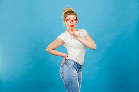 Crazy woman casual style nerdy glasses holding red fake lips on stick having fun, on blue. Photo take and carnival funny accessories concept.の写真素材