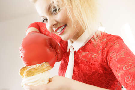 Dieting, weight loss concept. Funny blonde woman holding yummy choux puff cake with whipped cream and boxing glove, fighting off bad food. On greyの写真素材