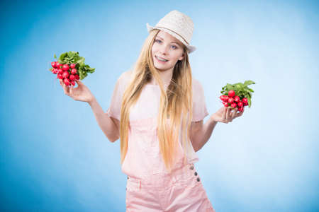 Positive teenage blonde long hair girl wearing summer clothing sun hat holding two bunches of fresh radish, on blueの写真素材