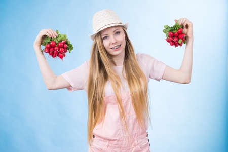 Positive teenage blonde long hair girl wearing summer clothing sun hat holding two bunches of fresh radish, on blueの写真素材