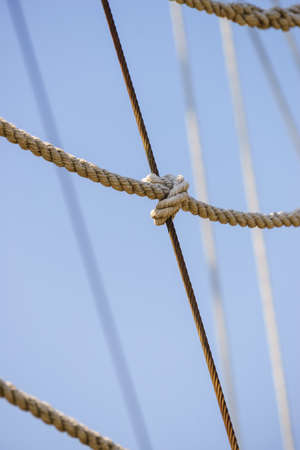 Old fashioned mast rigging, many brown ropes on clear blue sky background.