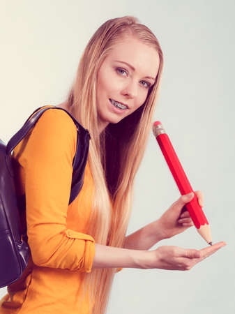 Happy young blonde teenage girl going to school or college wearing backpack and holding big pencil.の写真素材