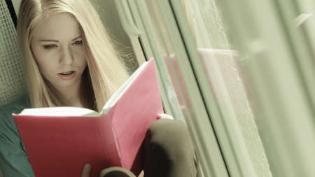 Young woman sitting on windowsill reading interesting book, having emotional face expression.の写真素材