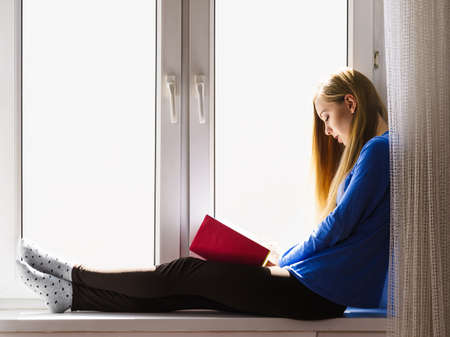 Leisure, literature and people concept. Young woman teen girl reading book at home while sitting on window sill. Calm and coziness.の写真素材