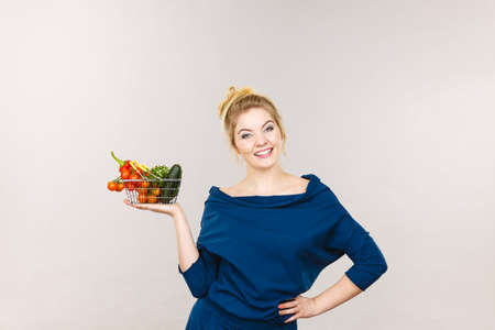 Buying good food, vegetarian products. Positive woman holding shopping basket with green red vegetables inside, recommending healthy high fibre diet, lifestyle modification, on greyの写真素材