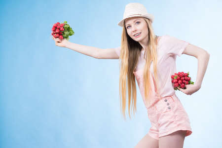 Positive teenage blonde long hair girl wearing summer clothing sun hat holding two bunches of fresh...