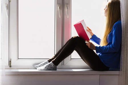 Leisure, literature and people concept. Young woman teen girl reading book at home while sitting on window sill. Calm and coziness.の写真素材