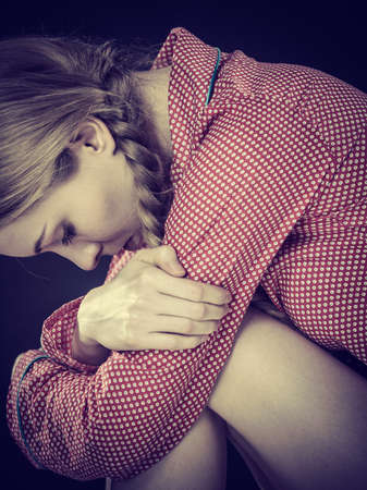 Mental health depression concept. Sad depressive young woman teen blonde girl wearing red pajamas sitting with arms around knee, on dark black wall backgroundの写真素材