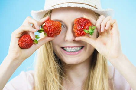 Young positive woman playing with fresh strawberries fruits, on blue. Healthy meal.の写真素材