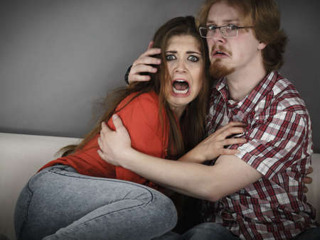 Funny couple sitting on sofa watching horror movie being scared hugging each other.の写真素材