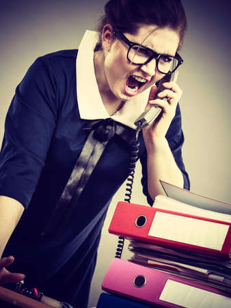 Mobbing at work, bad job relations concept. Angry mad bossy businesswoman talking on phone sitting working at desk full off documents in binders.の写真素材