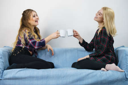 Two female friends sitting together on sofa chatting talking and drinking tea or coffee having fun spending great time.の写真素材
