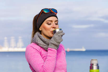 Woman athlete fit girl wearing warm sporty clothes outdoor in cold weather drinking hot tea from vacuum flask thermos, warming up. Sports, activities, hiking in winter or autumn timeの写真素材