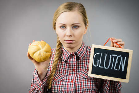 Young woman with braided hair holding small black board with gluten sign and bun bread roll. Bakery and bread allergy problem.の写真素材