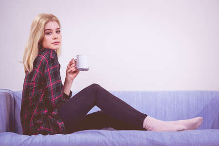 Pretty hipster young adult teenage woman sitting on sofa enjoying her leisure time drinking coffee or tea from mug.の写真素材
