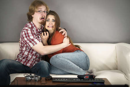 Funny couple sitting on sofa watching horror movie being scared hugging each other.