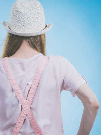 Unrecognizable woman back view. Female wearing cute outfit, pink dungarees, white tshirt and summer hat. Studio shot on blue backgroundの写真素材