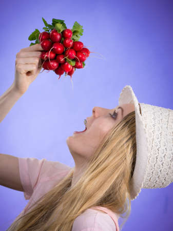 Positive teenage blonde long hair girl wearing summer clothing sun hat holding bunch of fresh radish, on violetの写真素材