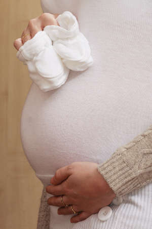 Pregnancy, motherhood concept. Pregnant woman holding little white baby socks and touching her...
