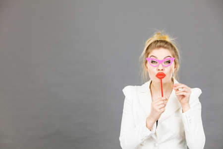 Happy elegant woman holding carnival accessories on stick having fun at work wearing white office...