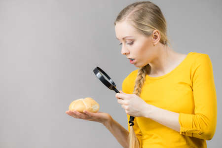 Shocked woman holding bun bread roll and magnifying glass examine wheat product ingredients seeing something disgustingの写真素材