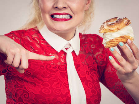 Skeptical woman holding choux puff with cream flavour having disgusted face expression being on diet. Unhealthy eating, sweet baked food concept.の写真素材