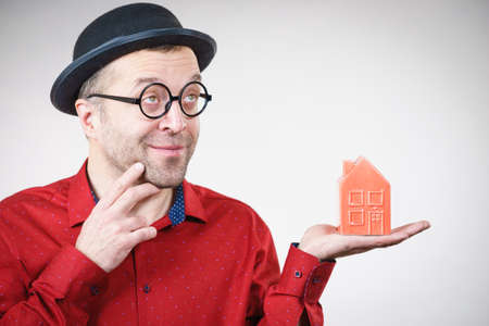 Adult man wearing funny hat and eyeglasses holding small red house model. Guy being real estate...