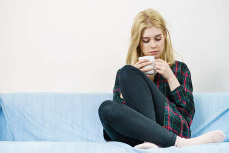 Sad contemplating woman sitting on couch alone drinking tea of coffee from mug.