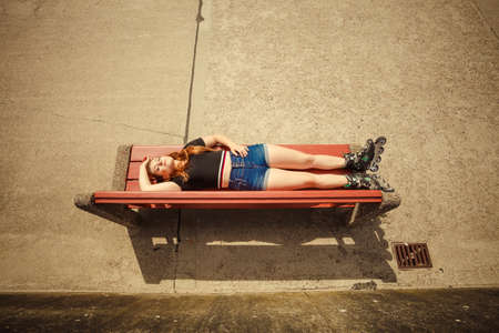 Happy joyful young woman wearing roller skates lying on bench with raised legs. Bizarre high angleの写真素材