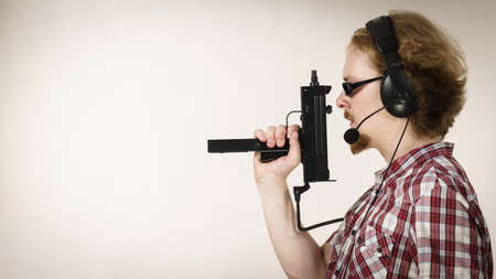Nerd geek young adult man playing on the video console holding gun wearing headphones with...