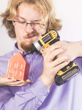 Man doing house renovation. Guy using drill on small red home model.